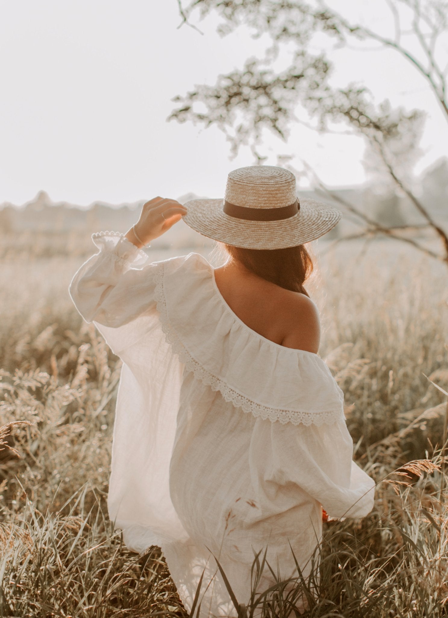 A woman in a flowing white dress and hat walks peacefully in a lush field, embracing the warmth of the sunny day