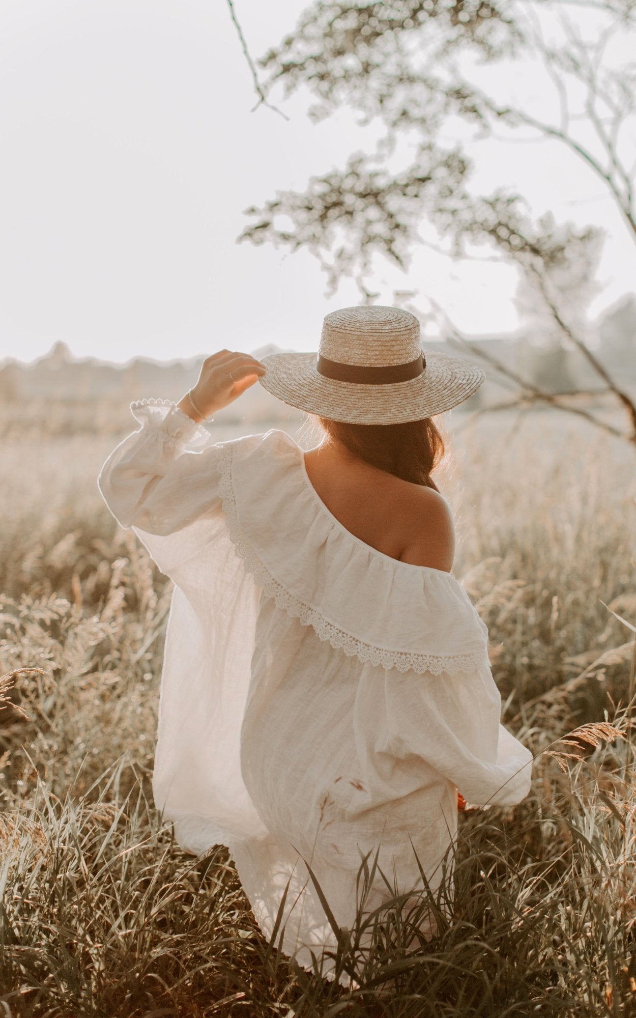 A woman in a flowing white dress and hat walks peacefully in a lush field, embracing the warmth of the sunny day